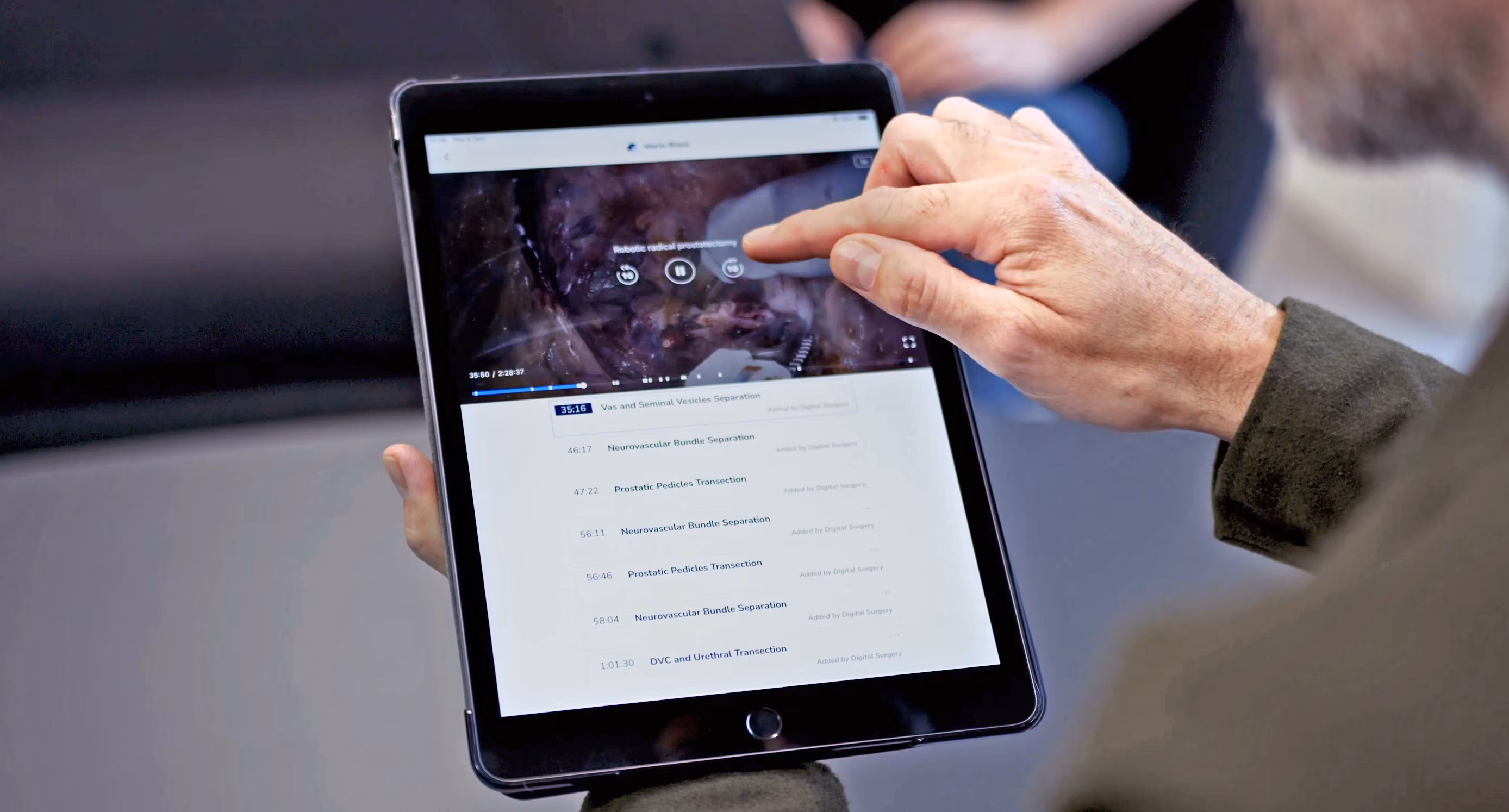 A surgeon scrubbing through a procedure on the Touch Surgery iPad app, with a list of timestamped chapter annotations sitting underneath the video